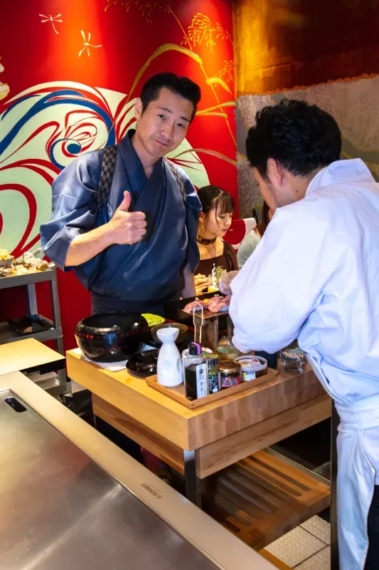 A sushi chef in a blue kimono giving a thumbs up at his restaurant counter