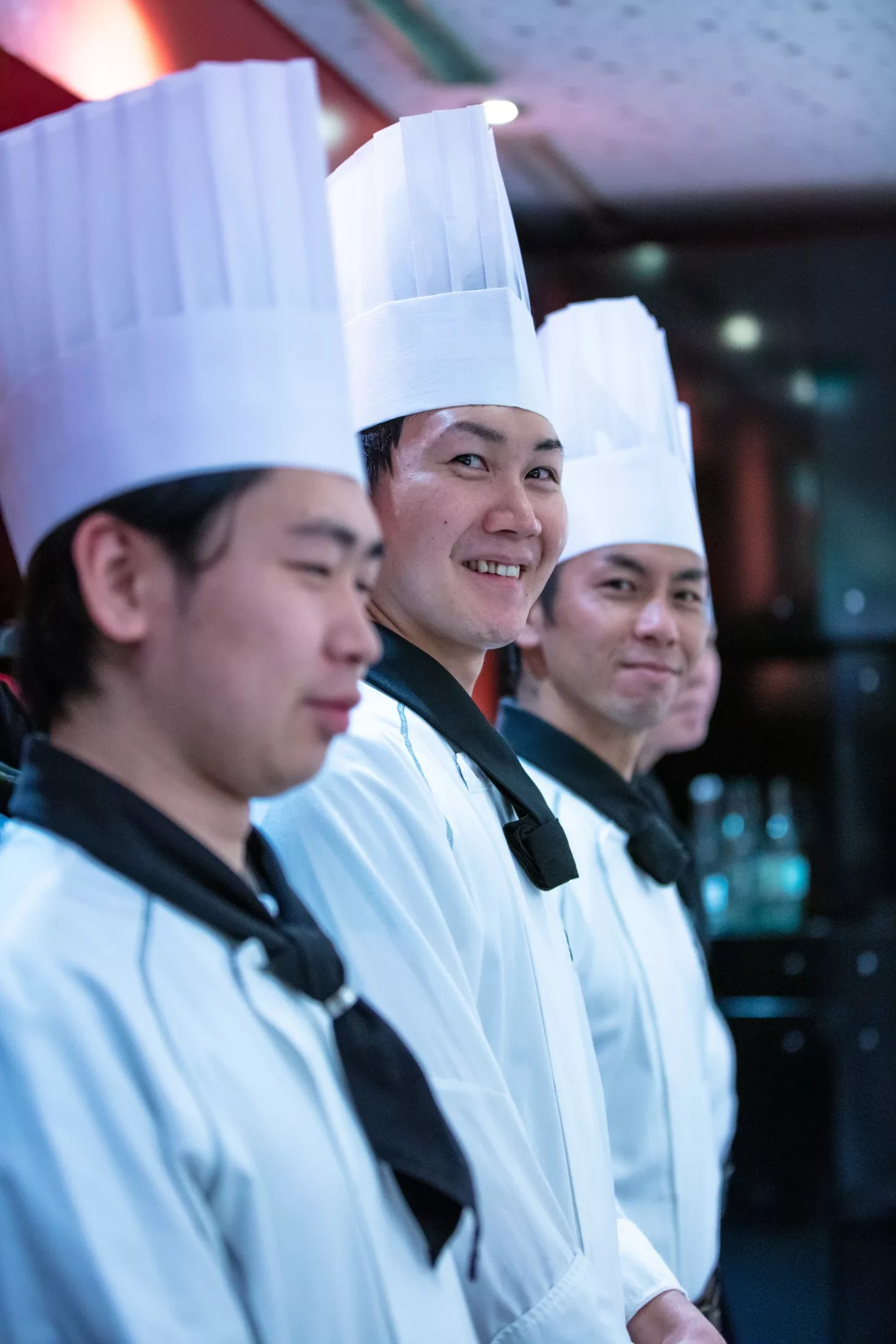 Three chefs of a Japanese teppanyaki kitchen in tall white toques, one turning to smile at the camera