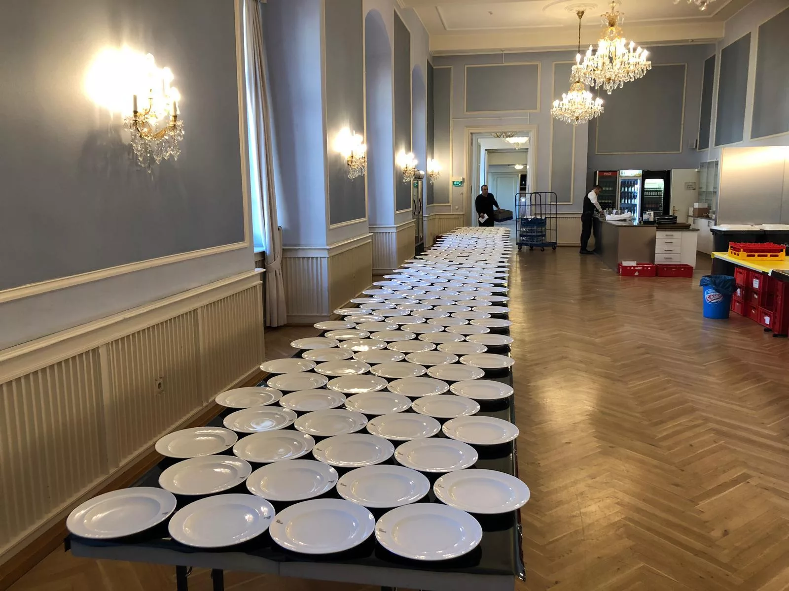 Centenas de pratos dispostos sobre longas mesas em um salão de banquetes com lustres, antes do serviço