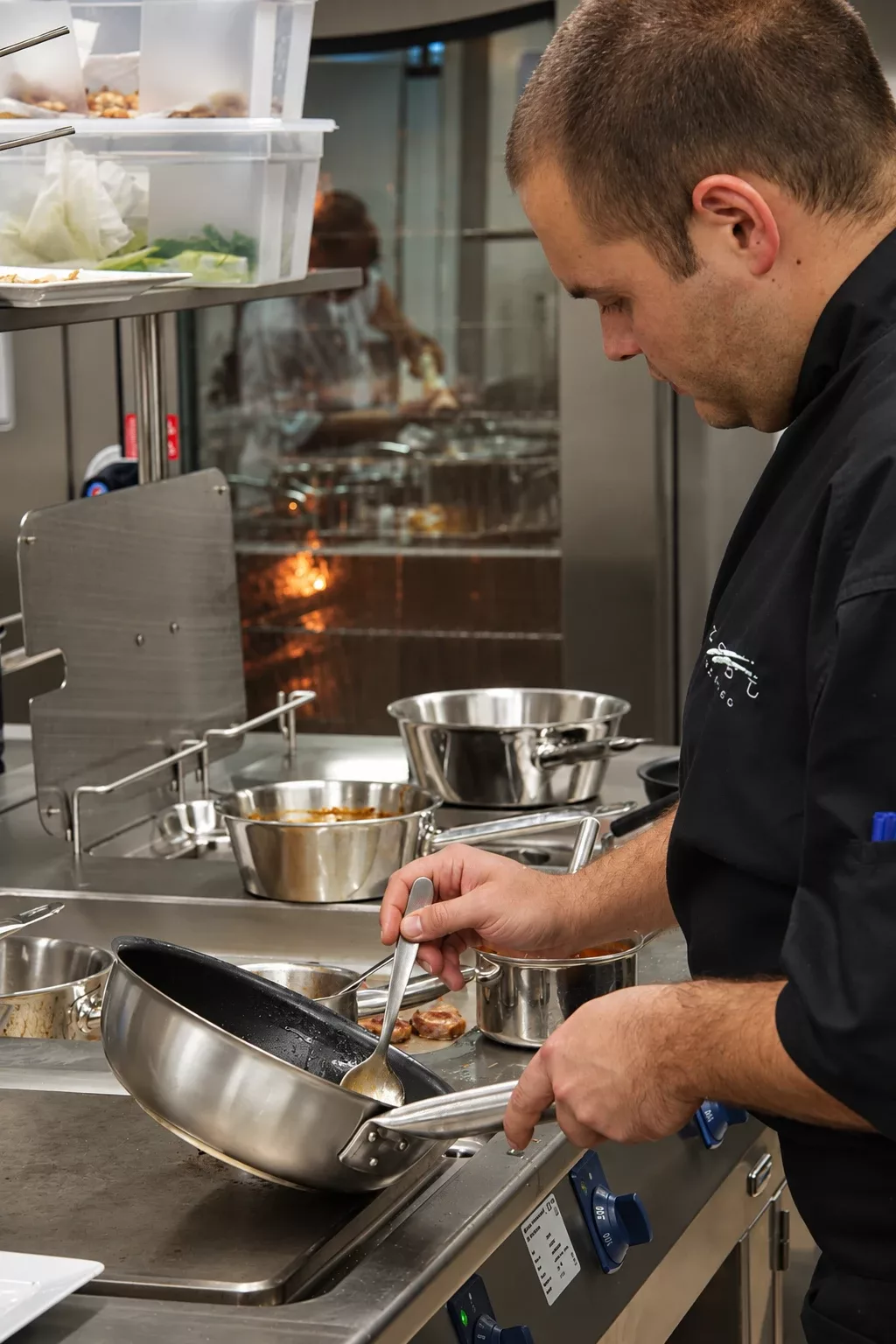 A sous chef in a black jacket on the hot line of the Monaco restaurant, spooning a sauce in a pan, with the pass behind him