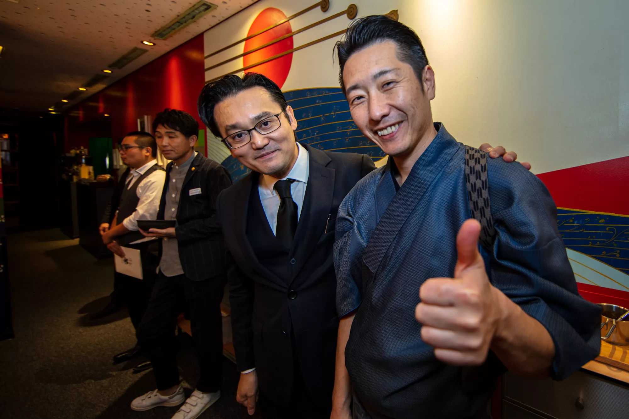 Two Japanese restaurant hosts smiling at an evening tasting, one in a suit, one in a blue kimono giving a thumbs up